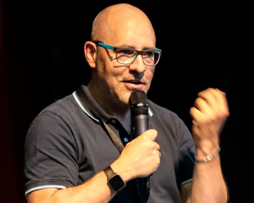 Zu sehen ist Matthias Blaß. Er trägt ein dunkles Poloshirt und hält ein Mikrofon vor den Mund. Er trägt eine blaue Brille und hat sehr kurze Haare. Er hat die linke Hand zu Faust geballt.