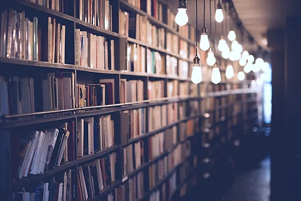 Bild zeigt ein langes Regal mit Veröffentlichungen und Büchern. Nach hinten wird das Bild unscharf. Der Raum wird von hängenden Glühbirnen beleuchtet.