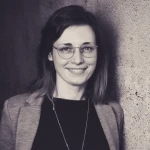 Profilbild von Ioana Reinke. Sie trägt eine Brille, ein dunkles Shirt und darüber einen Blazer.
