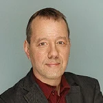 Profilbild von Rudolf Linsenbarth. Er trägt ein braunes Sakko und ein rotes Hemd. Er schaut freundlich in die Kamera.